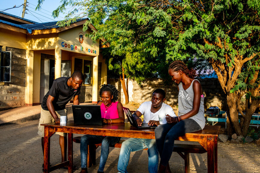 House in the background. In front four people. Two each look into a laptop together 